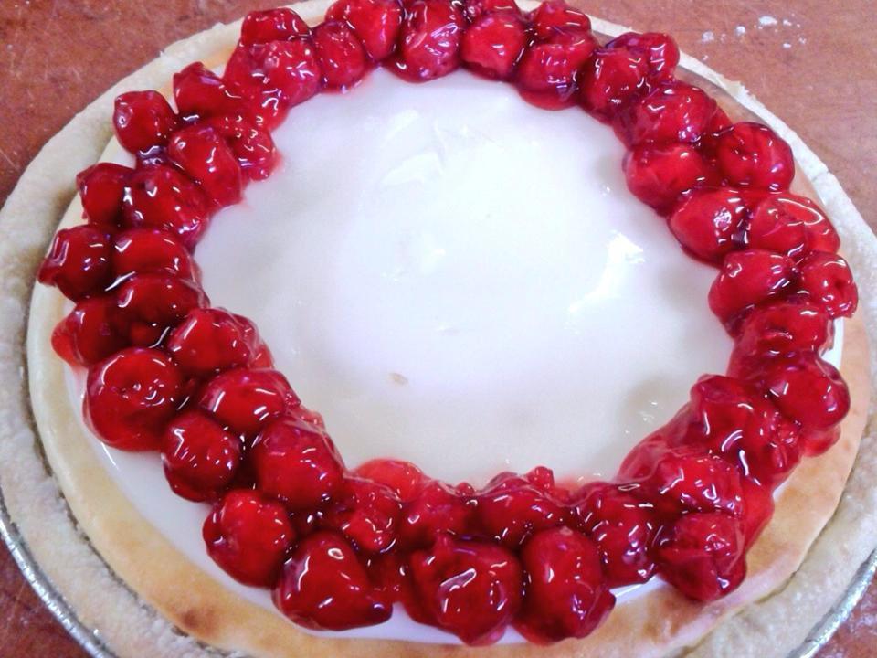 Close-up of a cream pie topped with a ring of glossy cherries on a floured wooden surface, red and white tones, appetizing